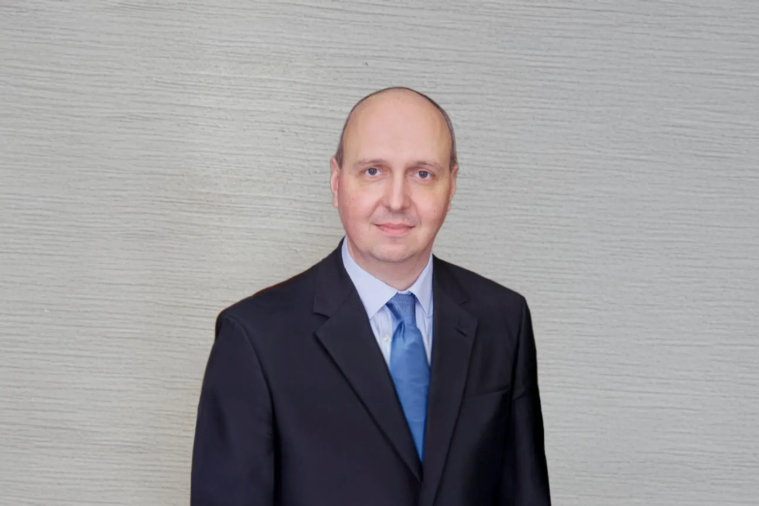 Person im dunklen Anzug und blauer Krawatte steht in einem Flur und lächelt leicht.