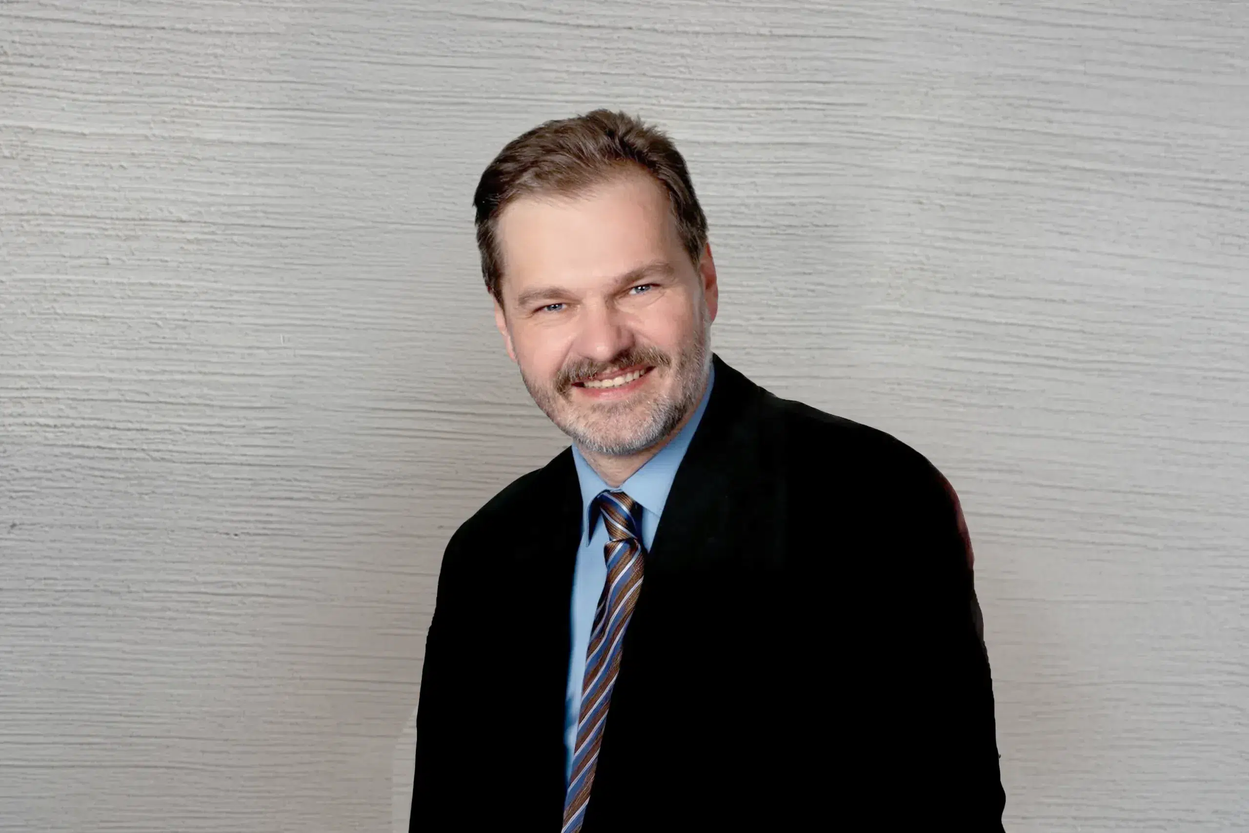 Lächelnde Person im Anzug mit blauem Hemd und gestreifter Krawatte vor grauem Hintergrund.