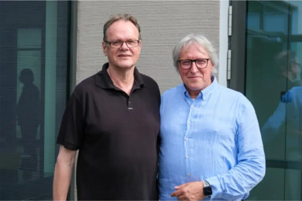 Zwei Männer posieren lächelnd vor einem Fenster, während im Hintergrund eine Silhouette zu sehen ist.