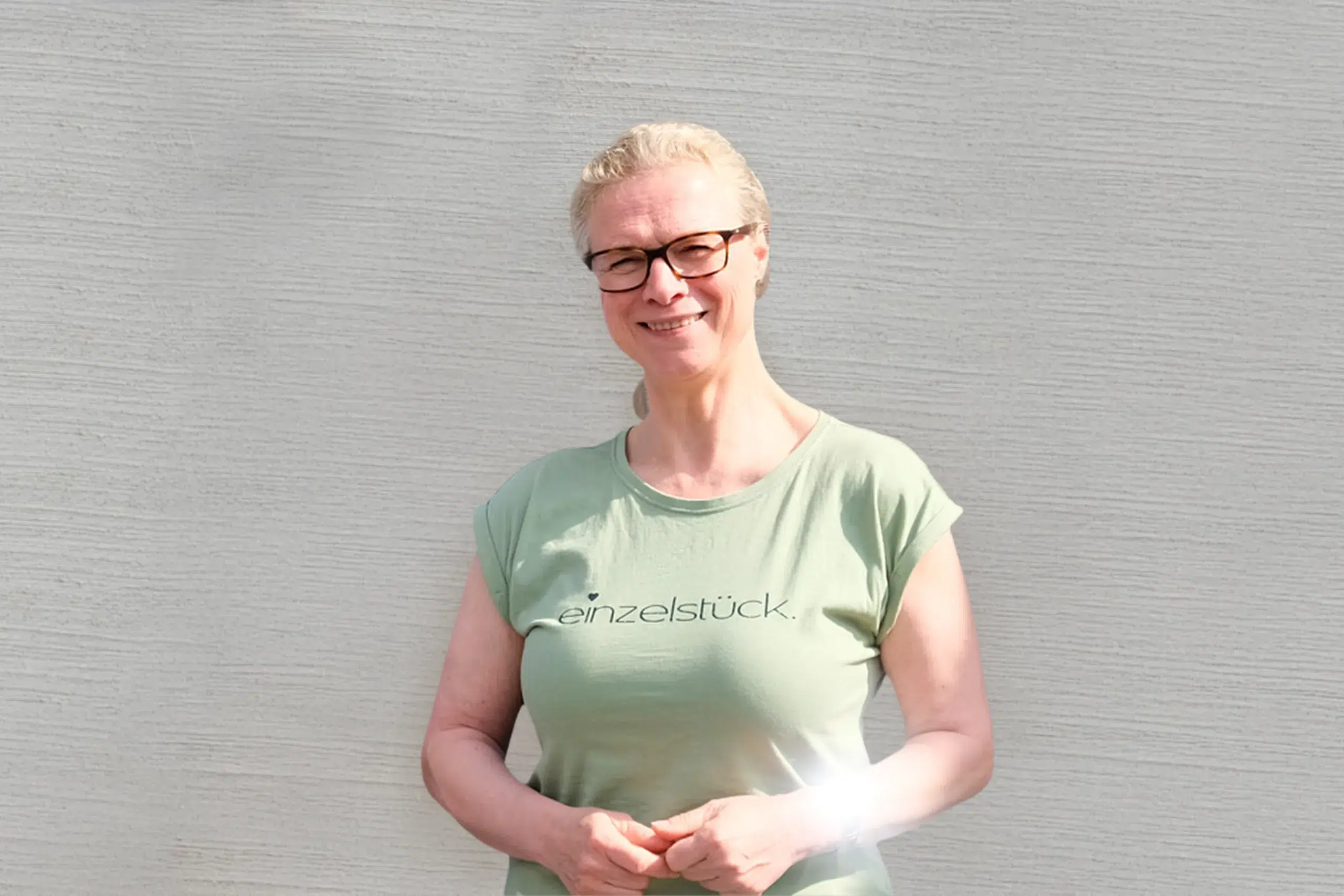Lächelnde Person vor heller Wand, trägt grünes T-Shirt mit Aufdruck ‚einzelstück‘.