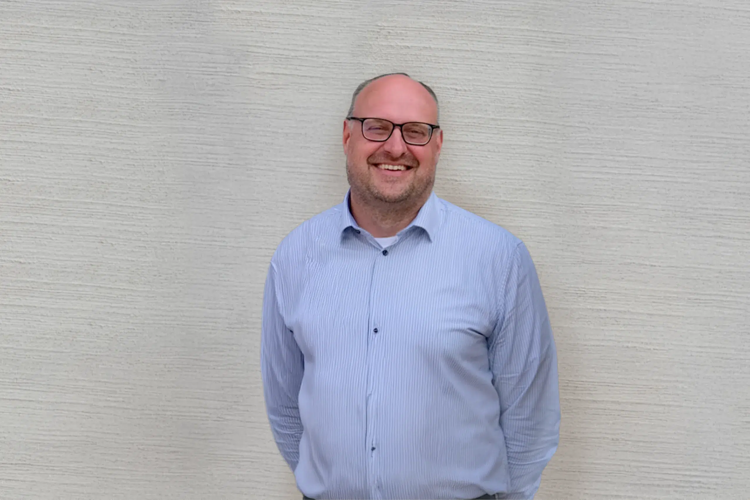 Mann mit Brille und Hemd steht lächelnd vor einer hellen Wand.