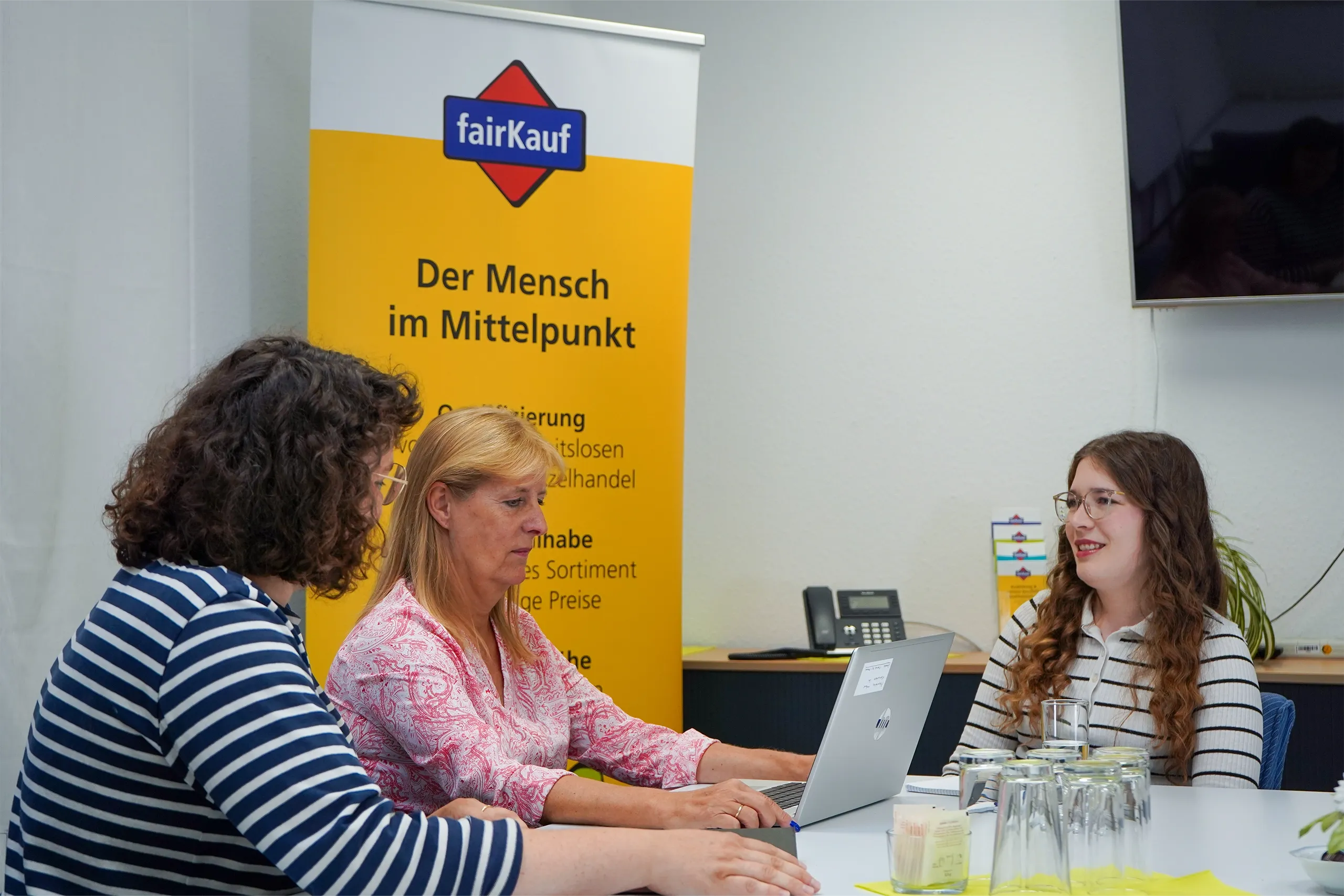Drei Personen sitzen im Meeting vor Laptop und fairKauf-Banner.