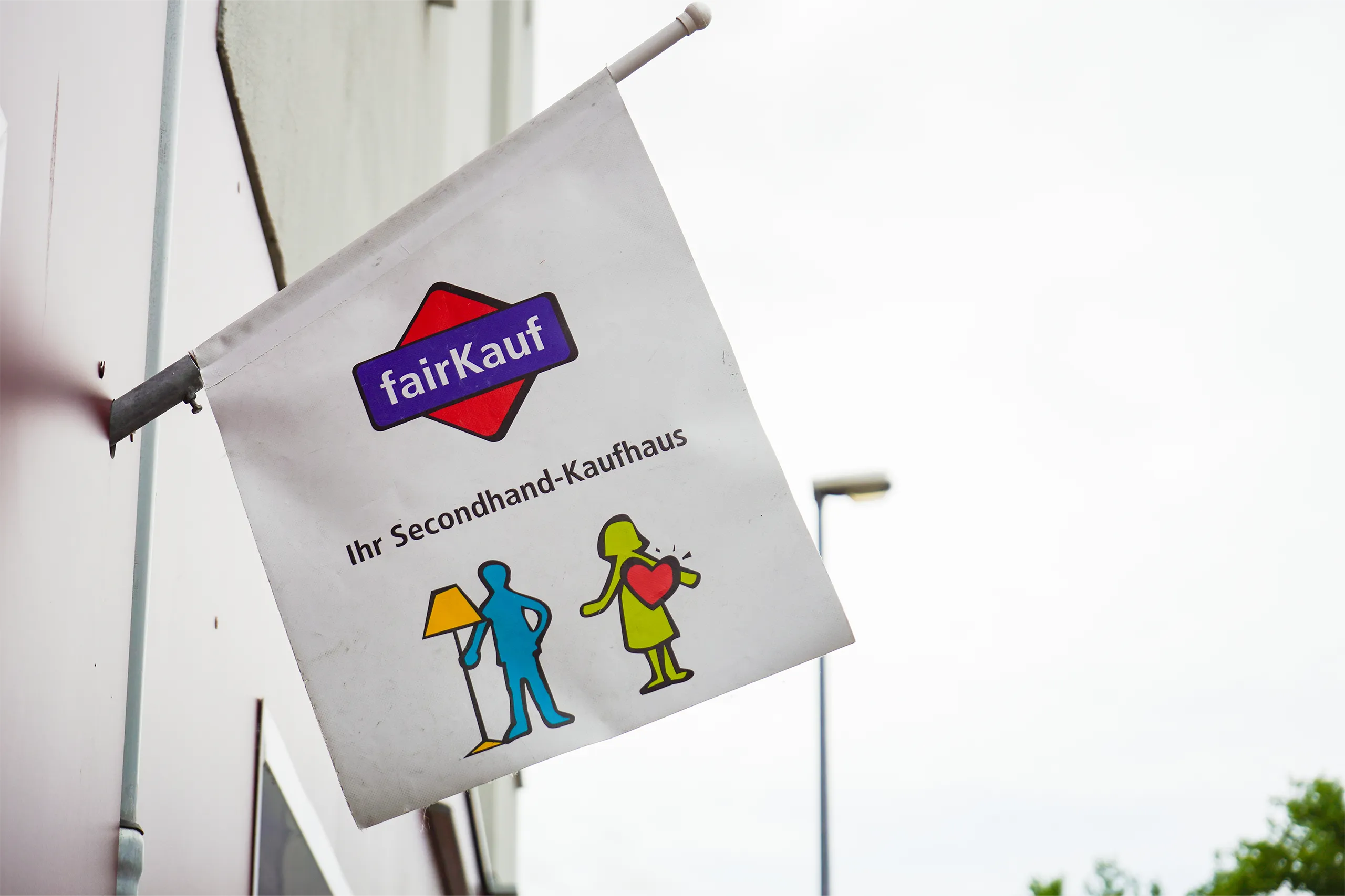 fairKauf-Fahne am Gebäude mit Logo und Figuren, die ein Secondhand-Kaufhaus bewerben.