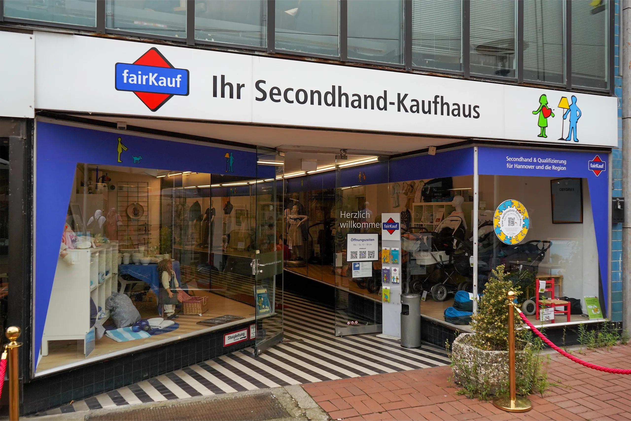 Eingang des fairKauf Secondhand-Kaufhauses mit großer Glasfront und Schaufenstern.