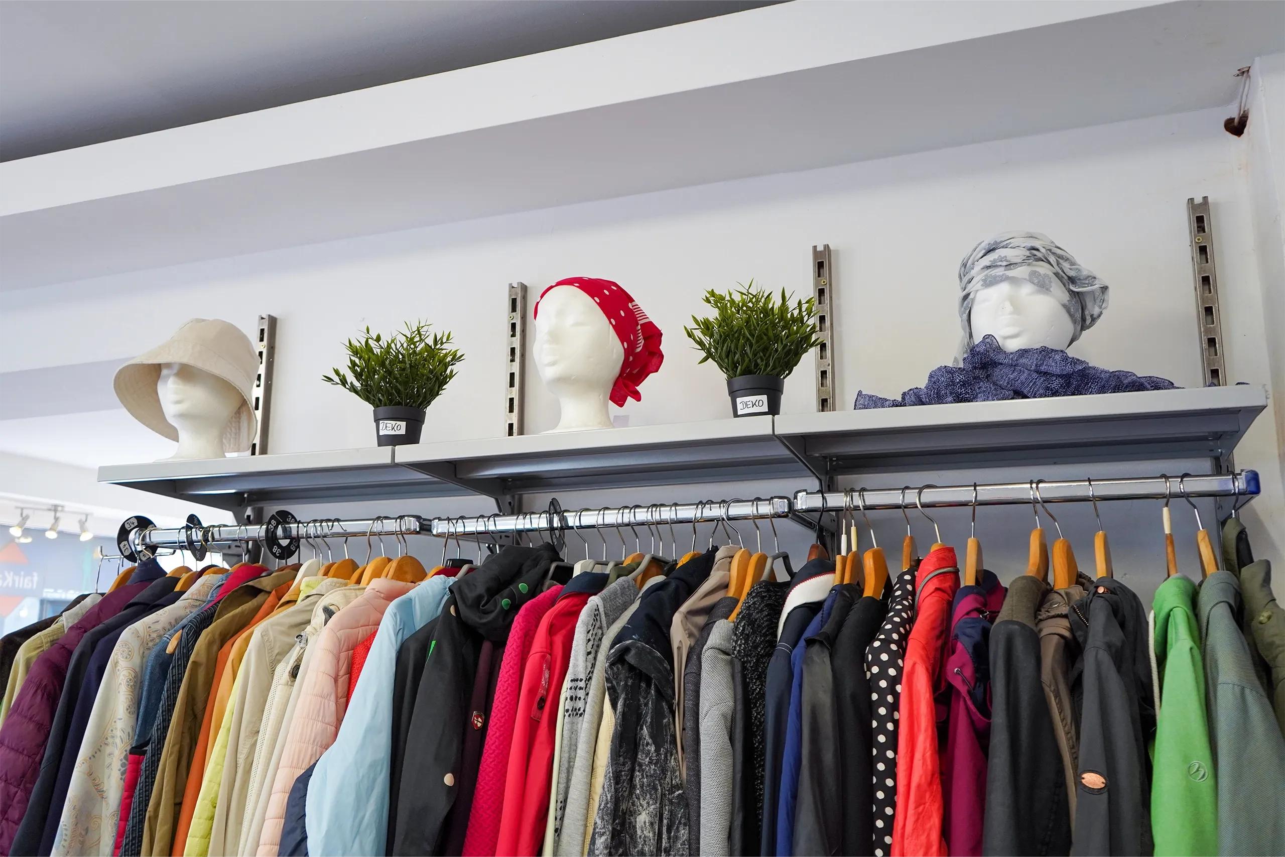 Kleiderstange mit bunten Jacken, oben Deko-Pflanzen und Hüte auf weißen Schaufensterköpfen.