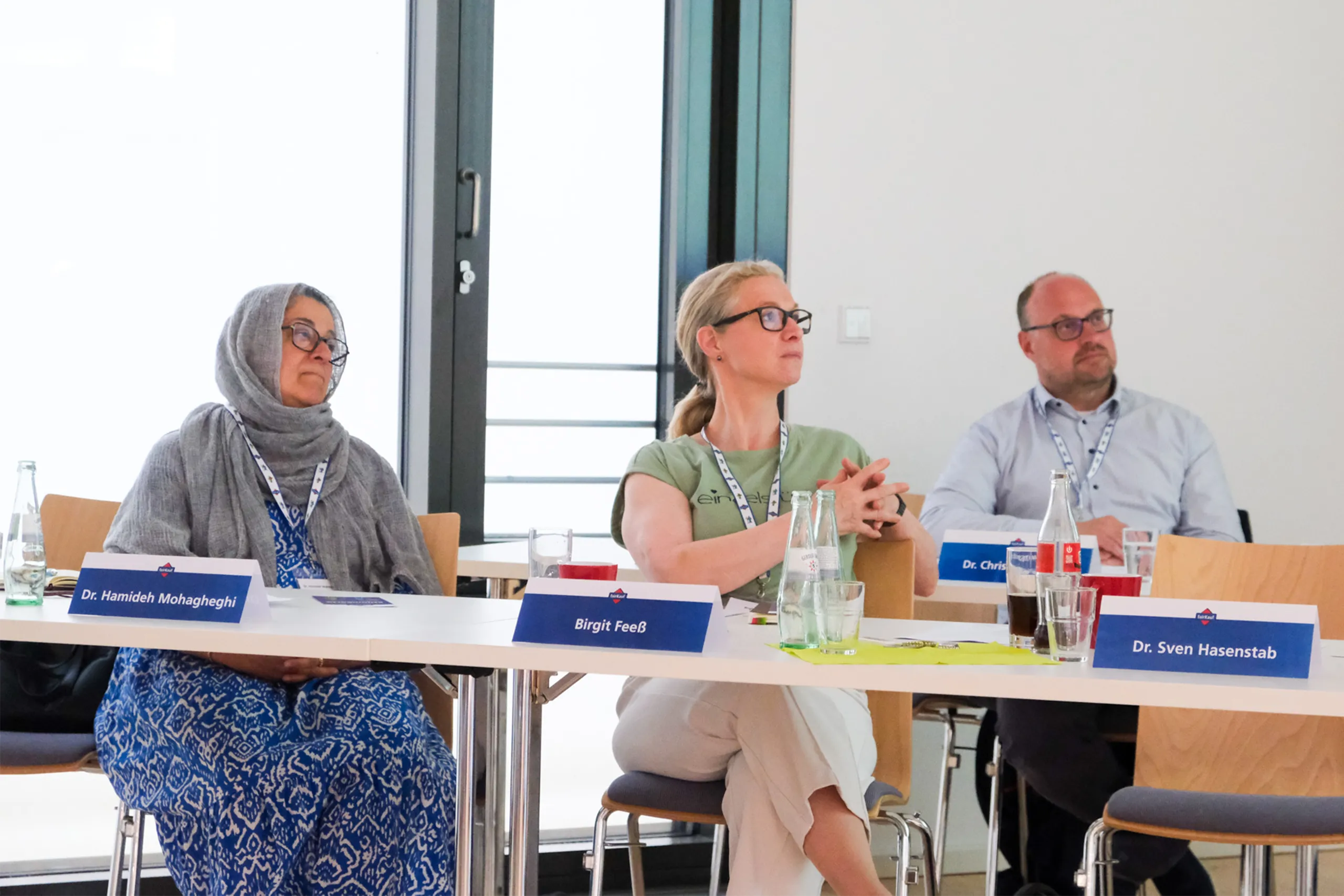 Drei Personen sitzen aufmerksam an einem Tisch mit Namensschildern in einem hellen Konferenzraum.