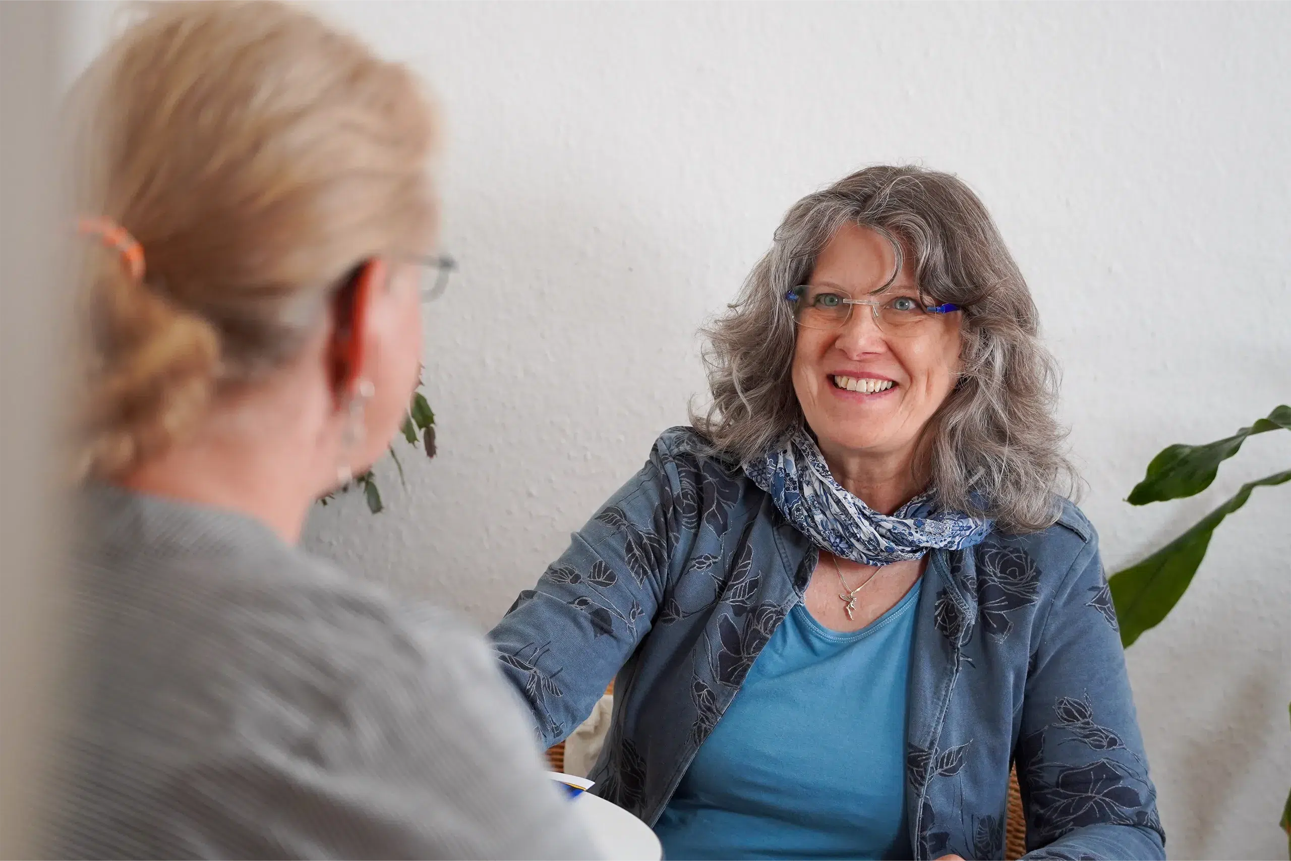 Zwei Frauen sitzen sich gegenüber; die Frau mit grauen Locken und Brille lächelt freundlich.