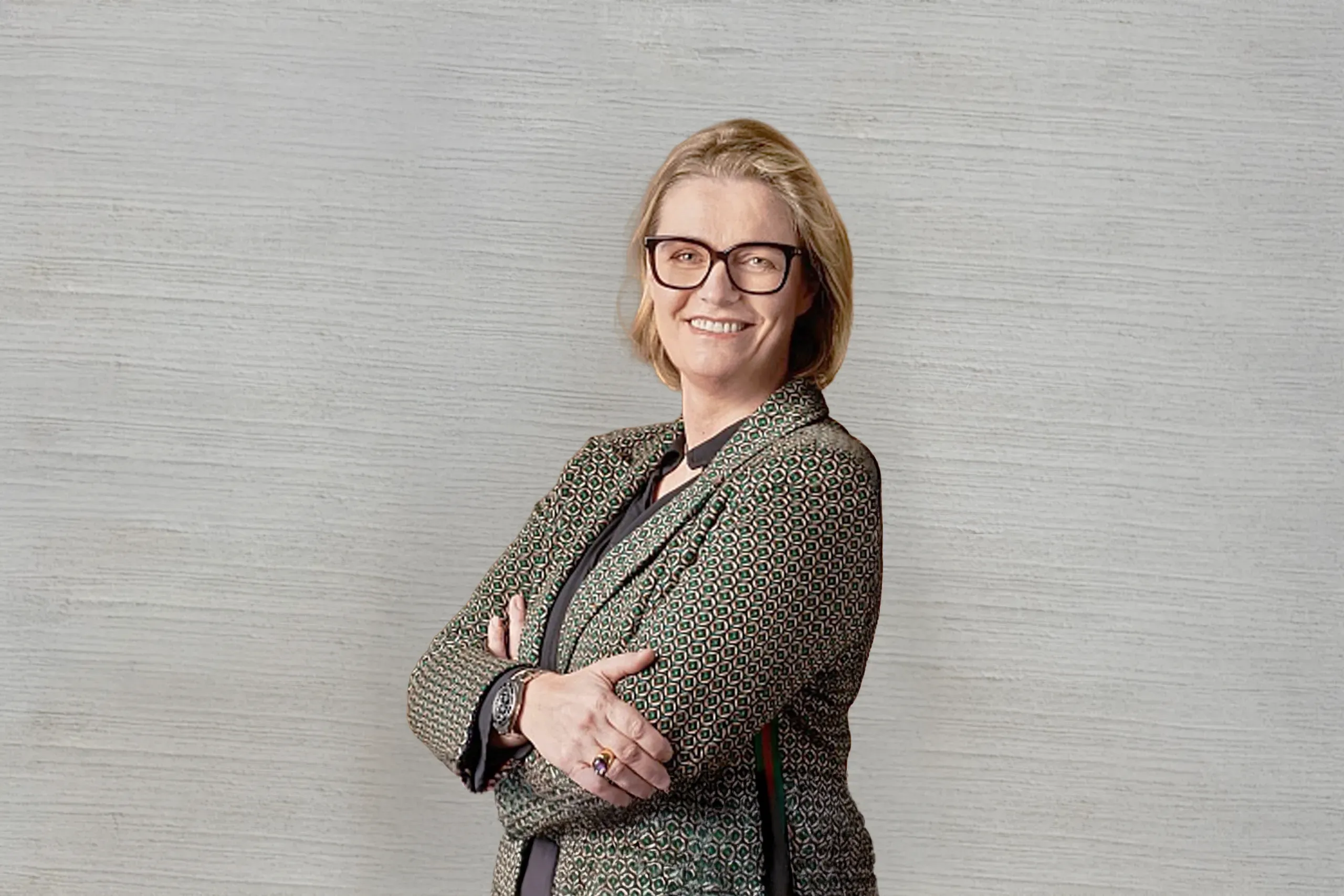 Frau mit Brille und gemusterter Jacke steht lächelnd vor heller Wand, Arme verschränkt.