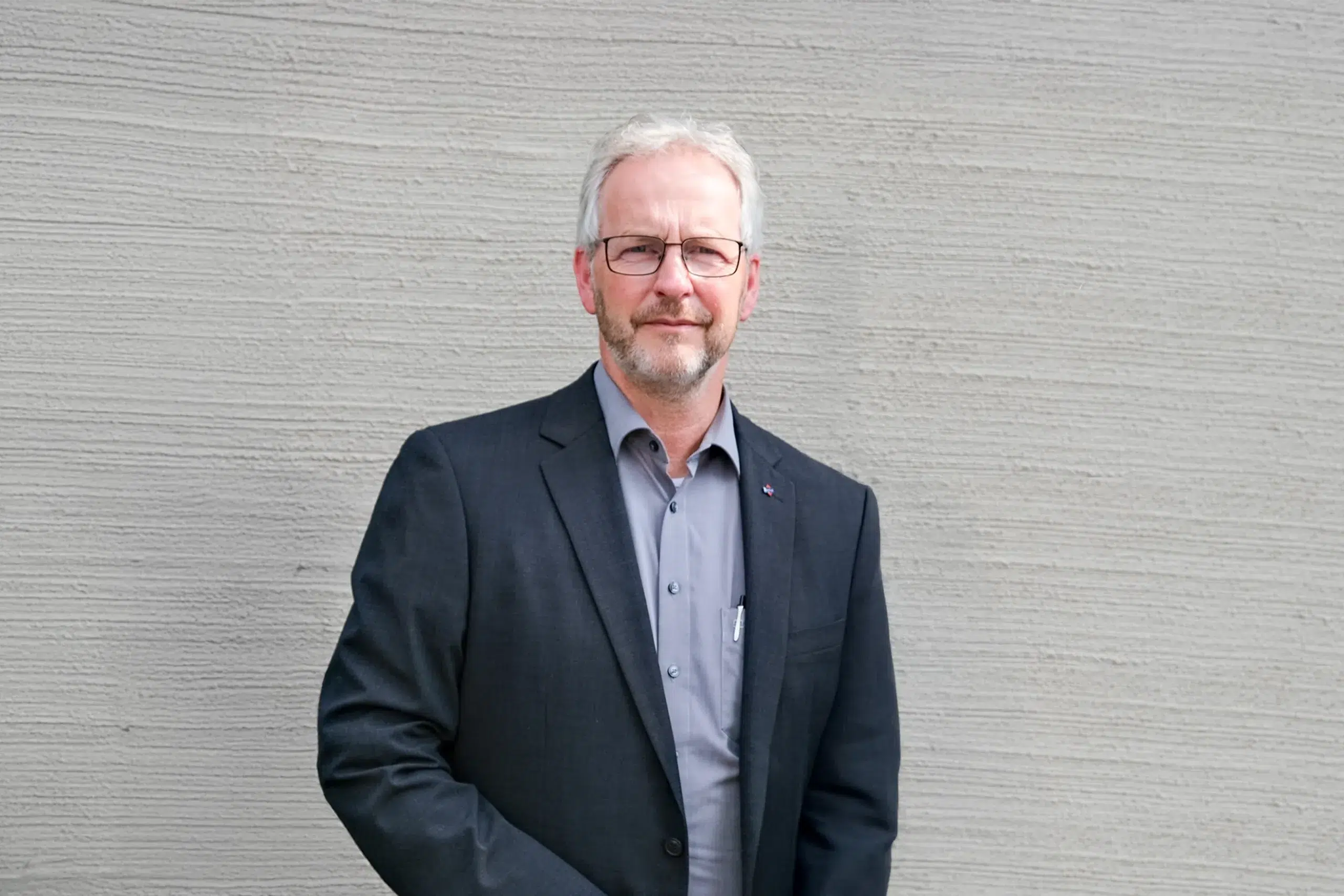 Mann im dunklen Anzug steht vor heller Wand und blickt ruhig in die Kamera.