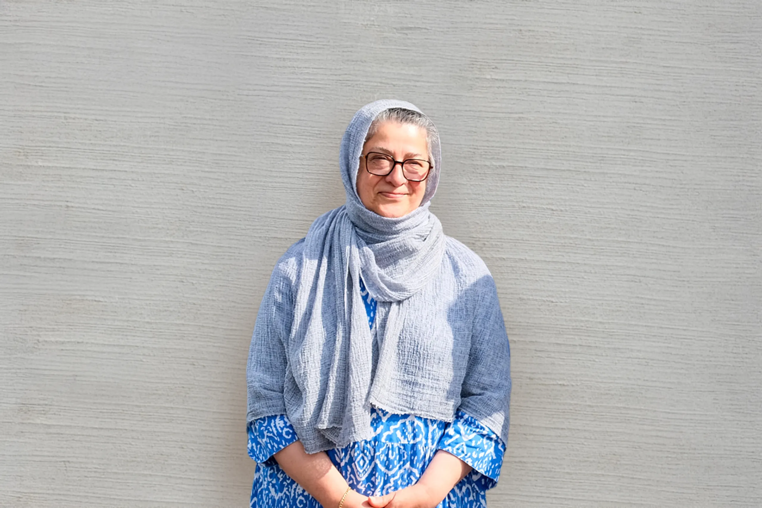 Lächelnde Frau mit grauem Kopftuch und blauem Kleid vor einer hellen strukturierten Wand.