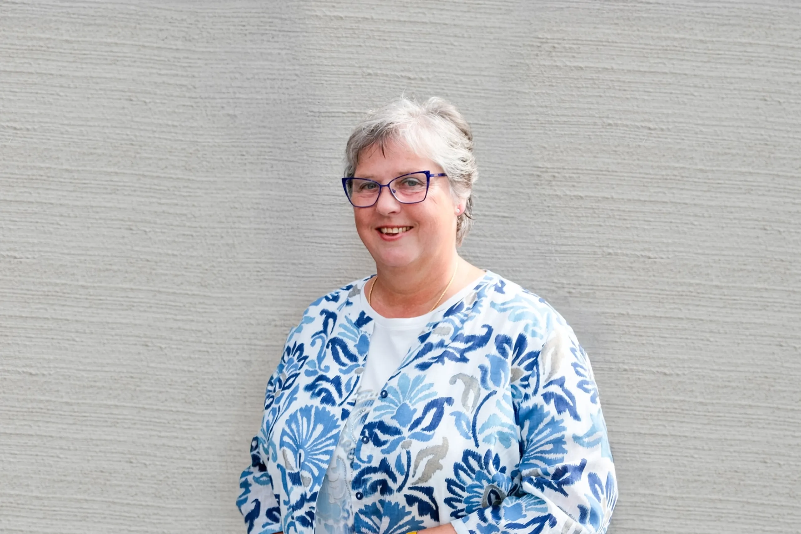 Lächelnde Frau mit blauer Brille und gemusterter Jacke vor heller strukturierter Wand.