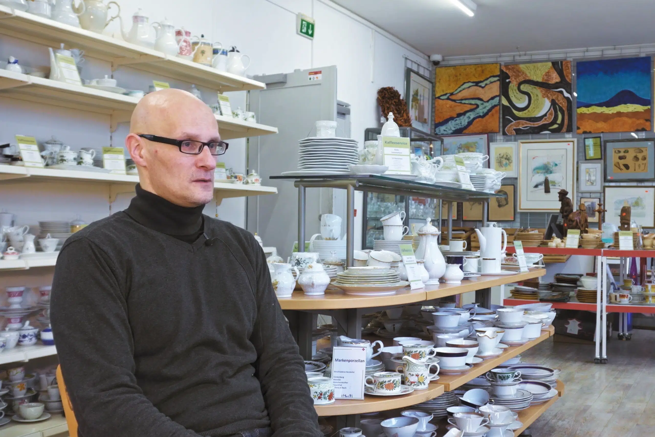 Mann sitzt im fairKauf-Geschirrbereich zwischen Regalen voller Porzellan und Keramik.