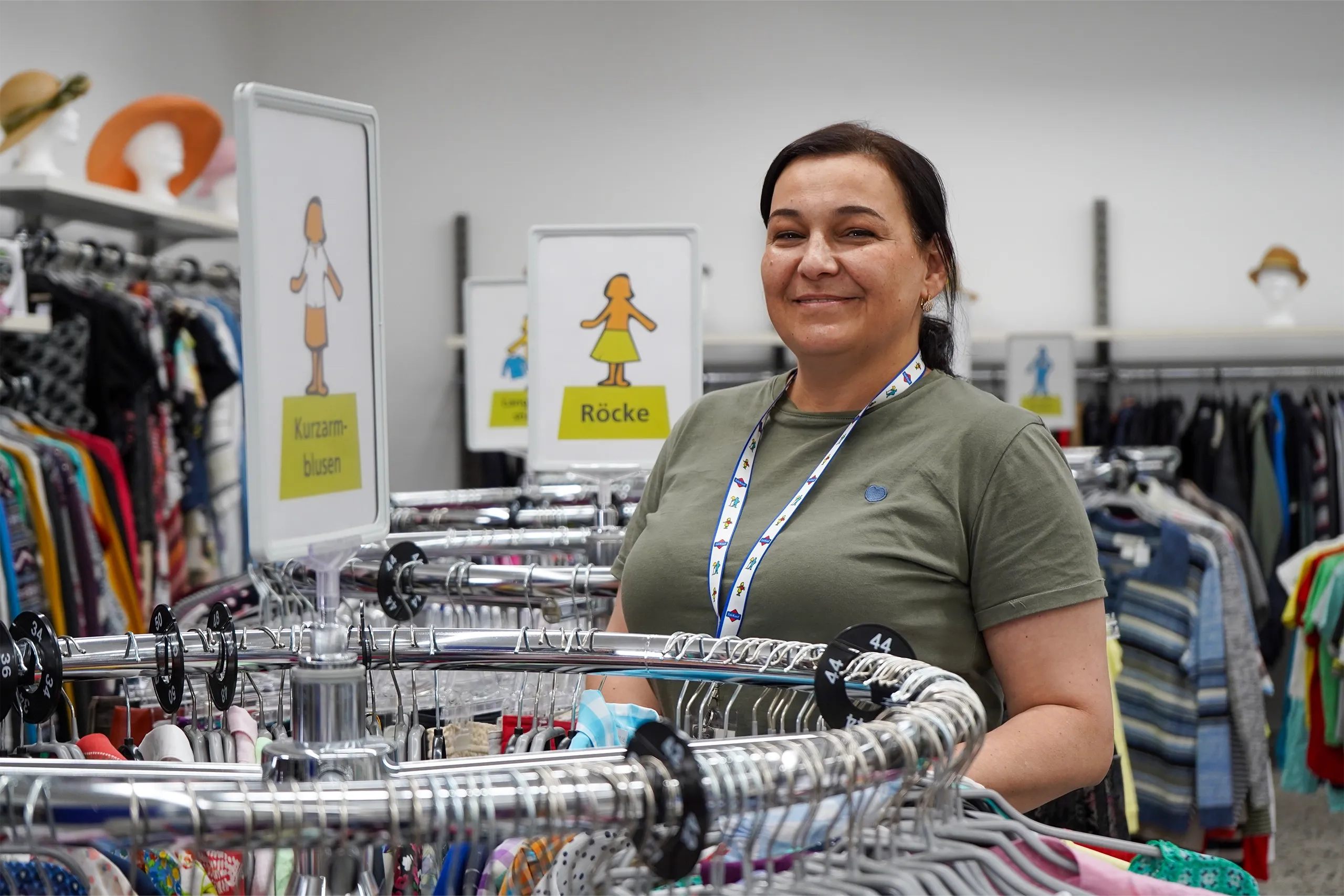 Eine lächelnde Frau in einem Geschäft für Secondhand-Kleidung steht neben Kleiderständern mit Kategorienchildern für Kurzarmblusen und Röcke.