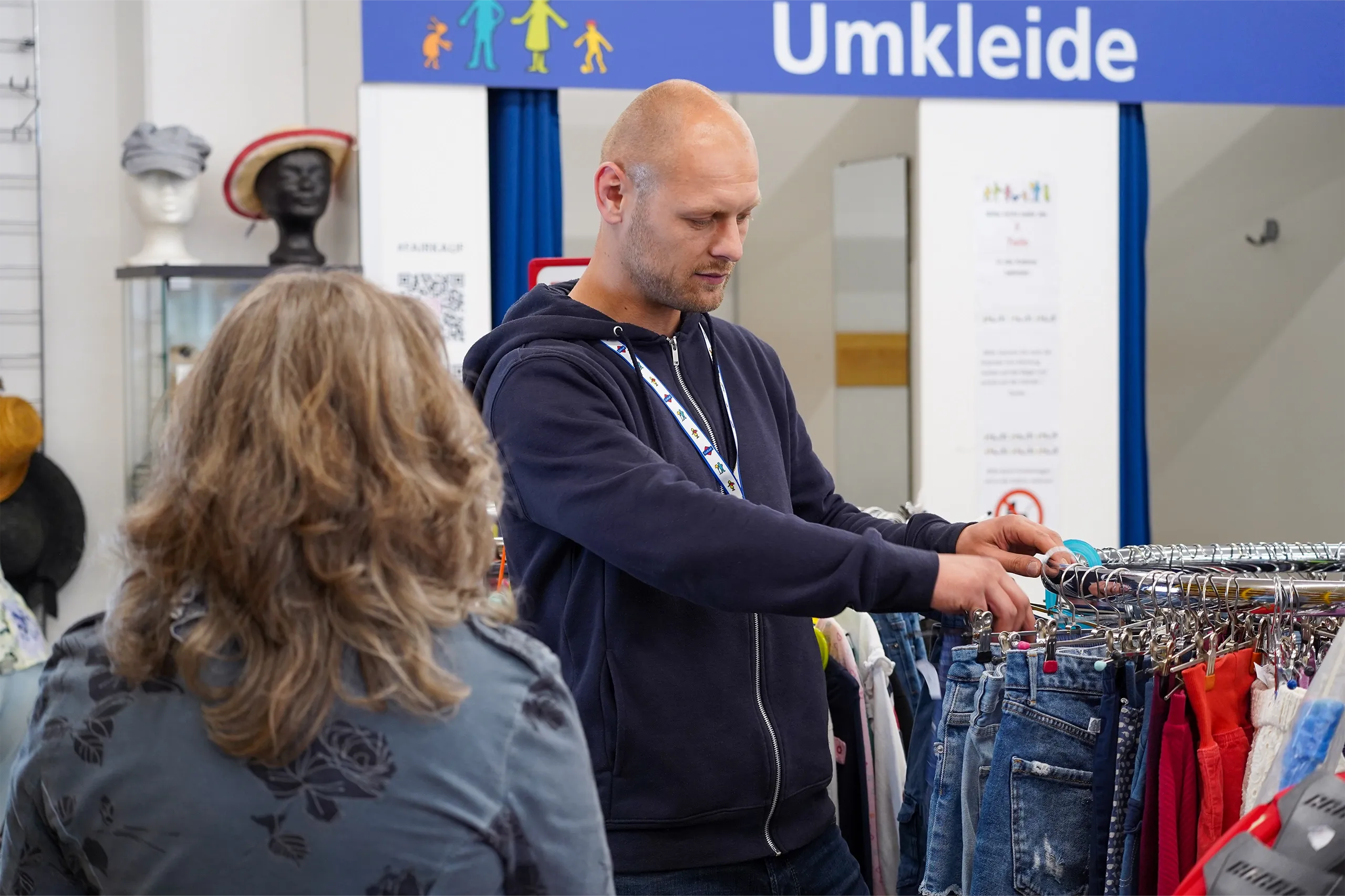 Ein Berater zeigt einer Kundin verschiedene Kleidungsstücke in einem Geschäft mit Umkleidebereich.