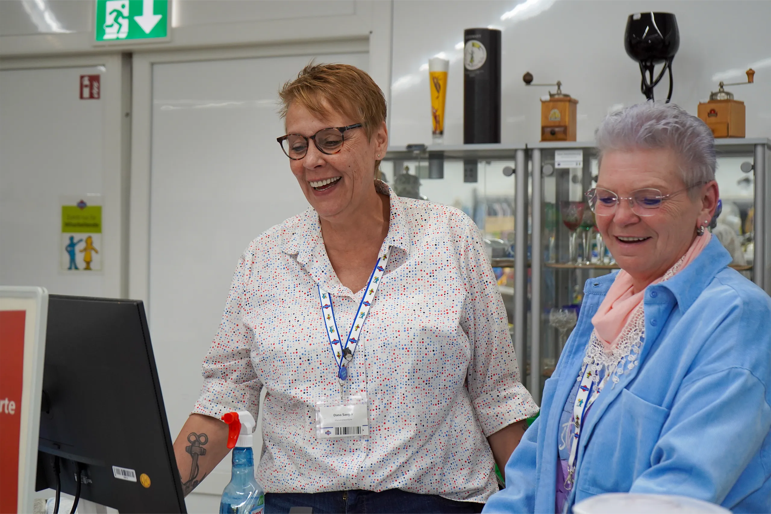 Zwei Frauen lächeln und arbeiten zusammen am Computer in einer hellen Umgebung.