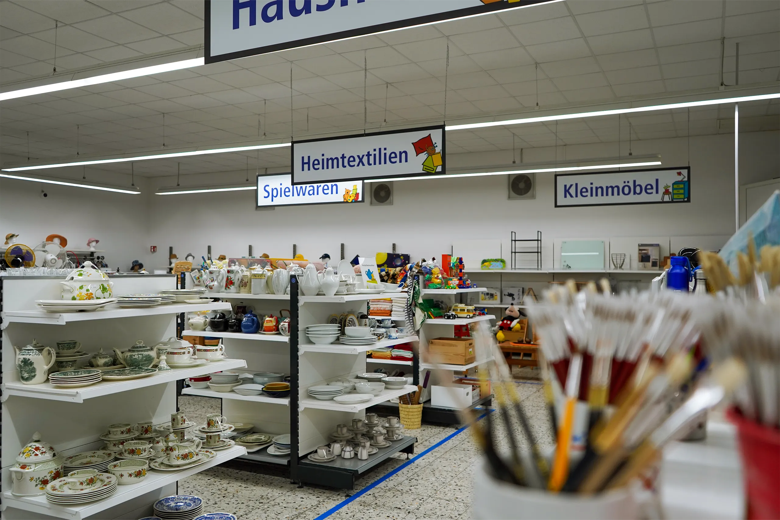 Regale mit Geschirr, Spielwaren und Heimtextilien in einem großzügigen Secondhandladen.