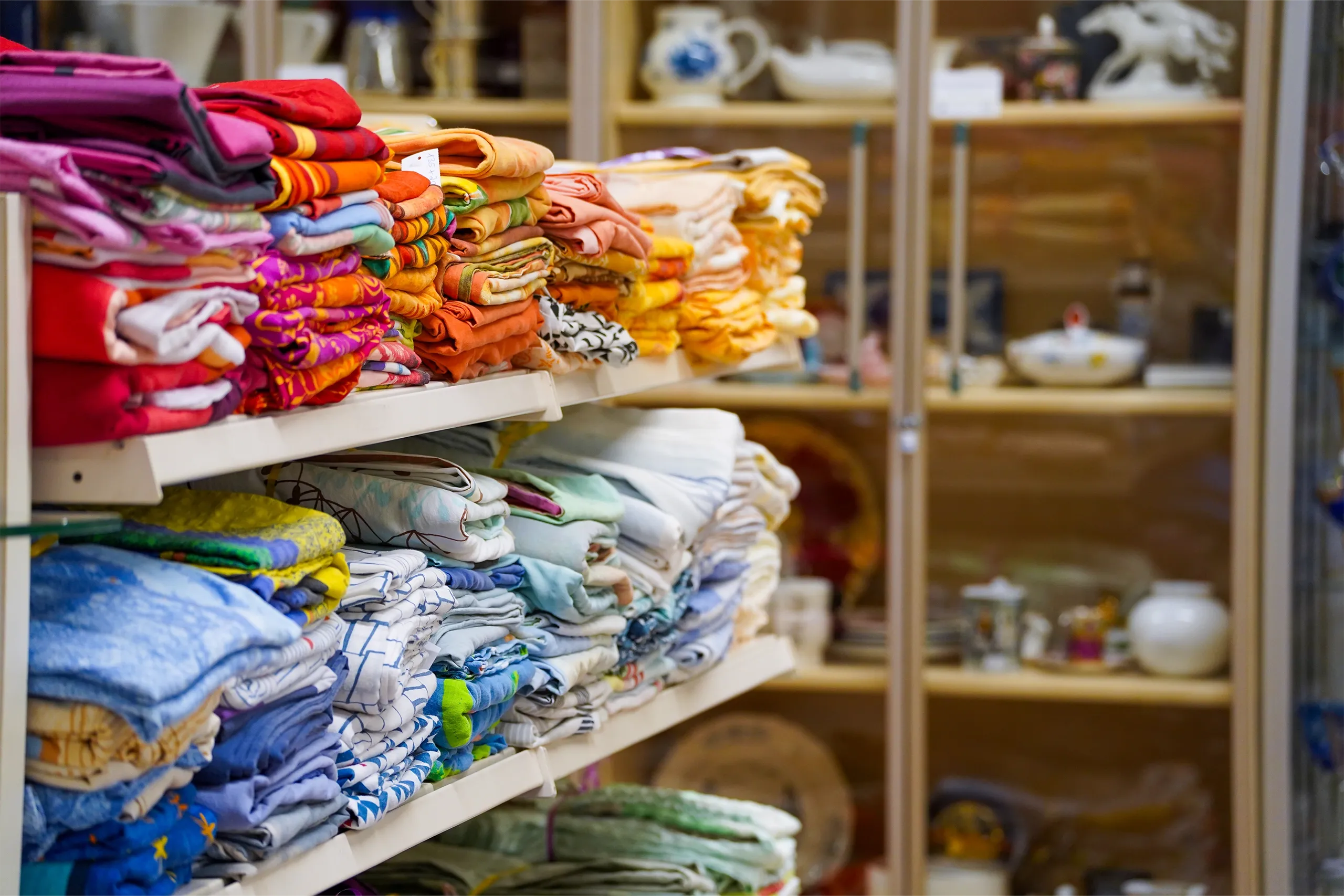 Gestapelte bunte Heimtextilien in Regalen, dahinter eine Glasvitrine mit Geschirr.