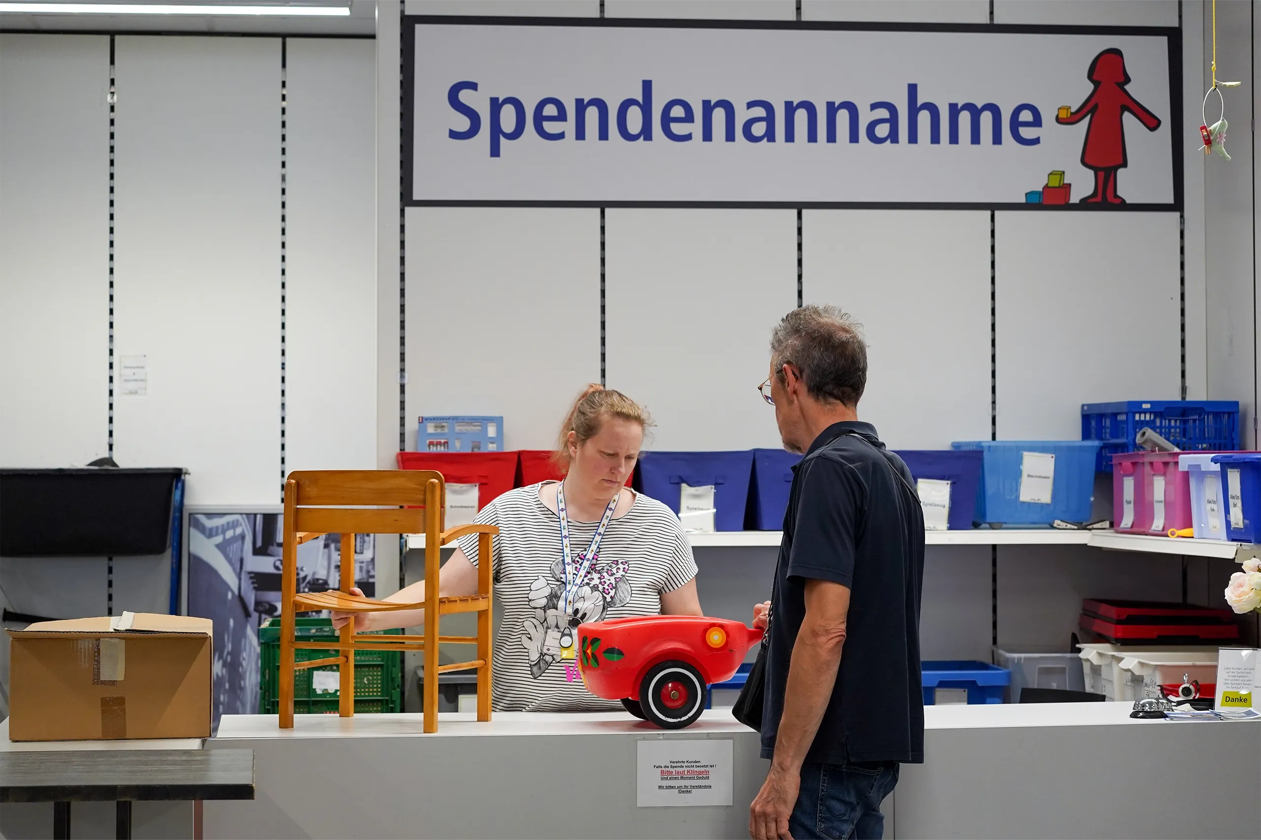 Eine Frau an einer Spendenannahme-Kasse spricht mit einem Mann, während ein rotes Spielzeugauto und ein Holzstuhl auf dem Tisch stehen.