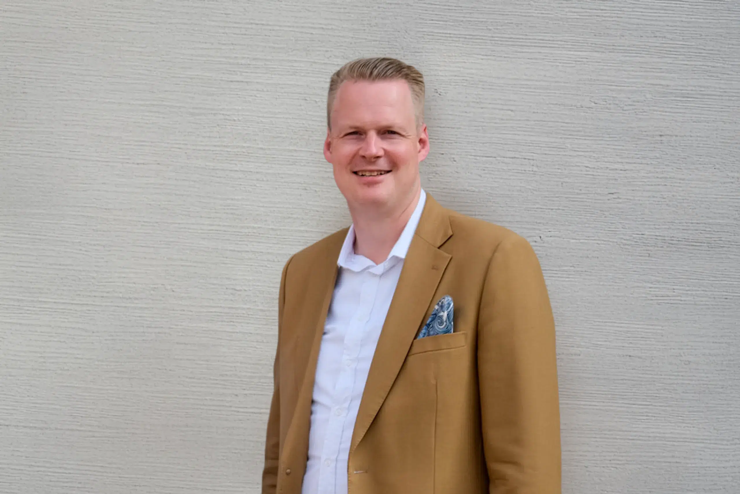 Lächelnder Mann im beigen Sakko und weißem Hemd steht vor einer hellen strukturierten Wand