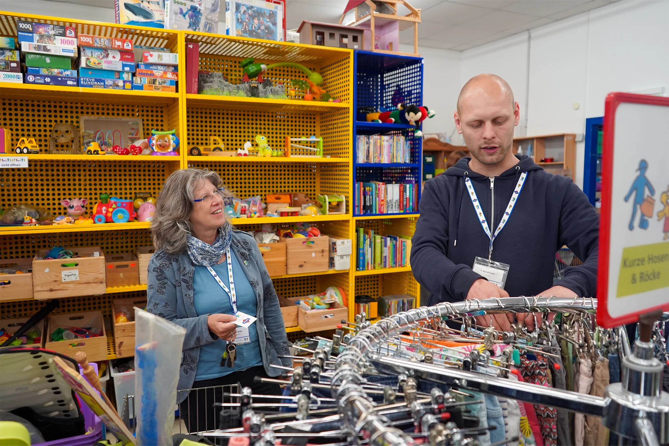 Zwei Mitarbeitende sortieren Kleidung in einer bunten Spiel- und Bücherabteilung.
