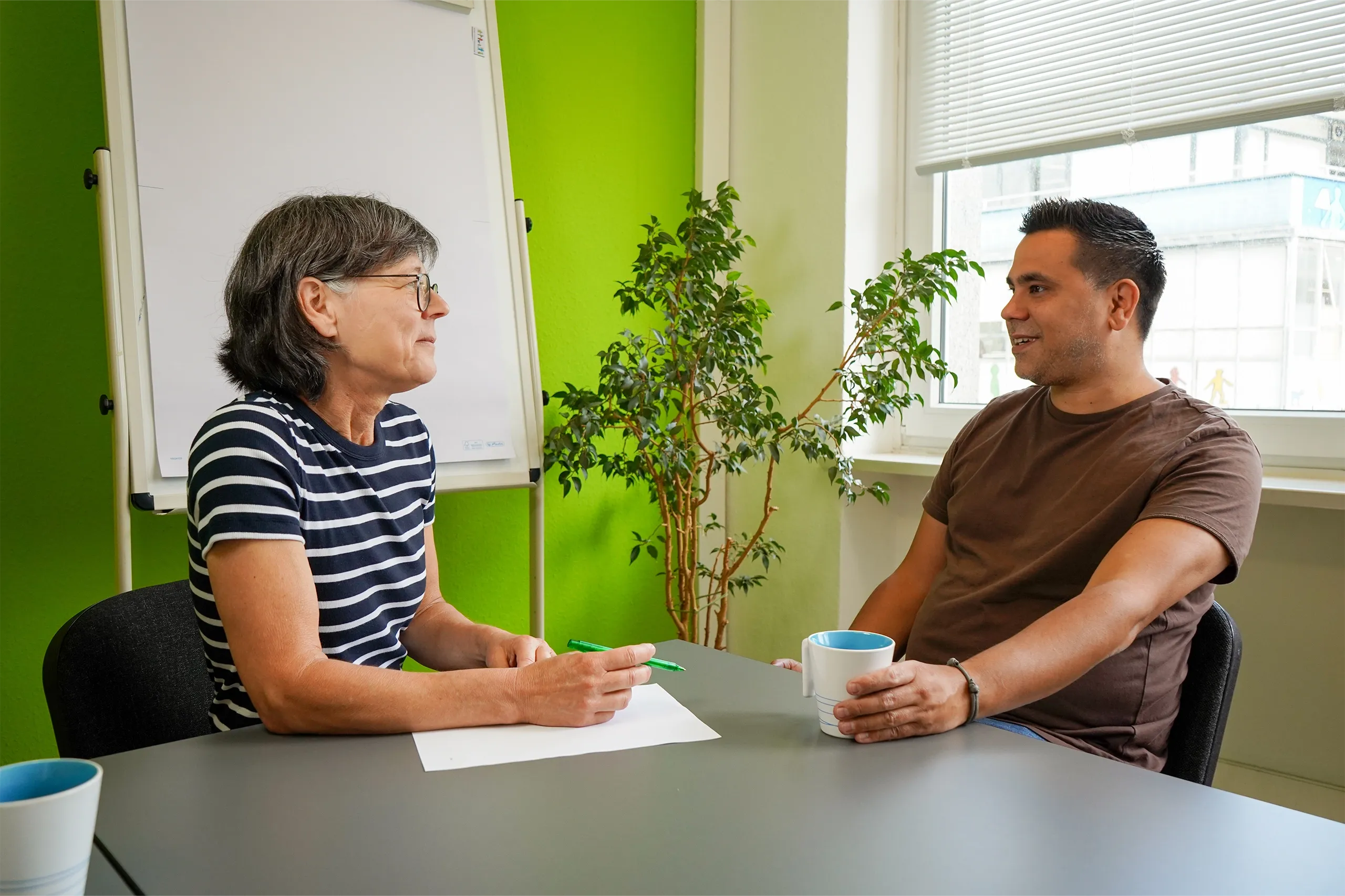 Zwei Personen im Beratungsgespräch an einem Tisch, mit Flipchart und Pflanze im Raum.