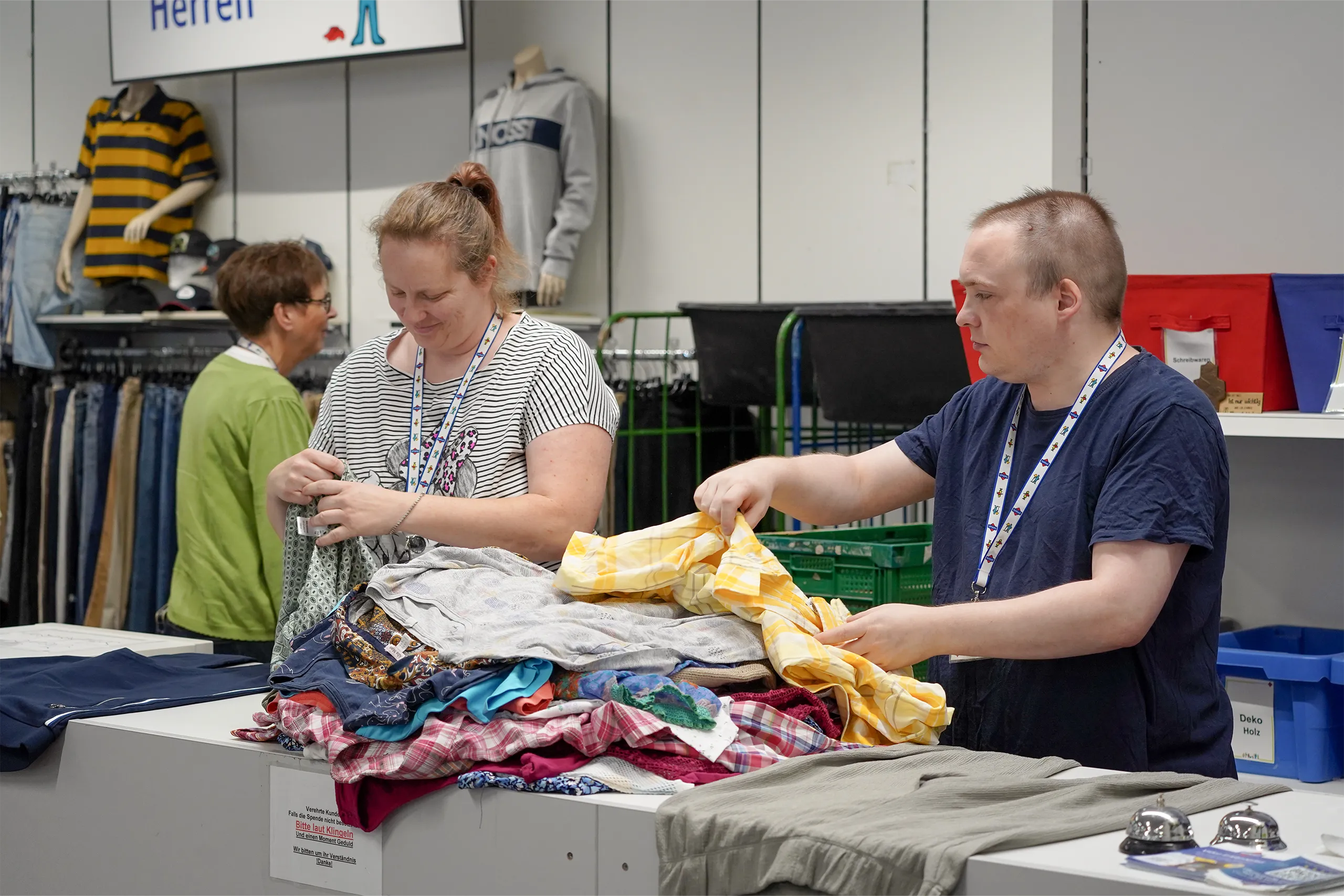 Zwei Personen sortieren Kleidung in einem Verkaufsraum mit verschiedenen Textilien im Hintergrund.