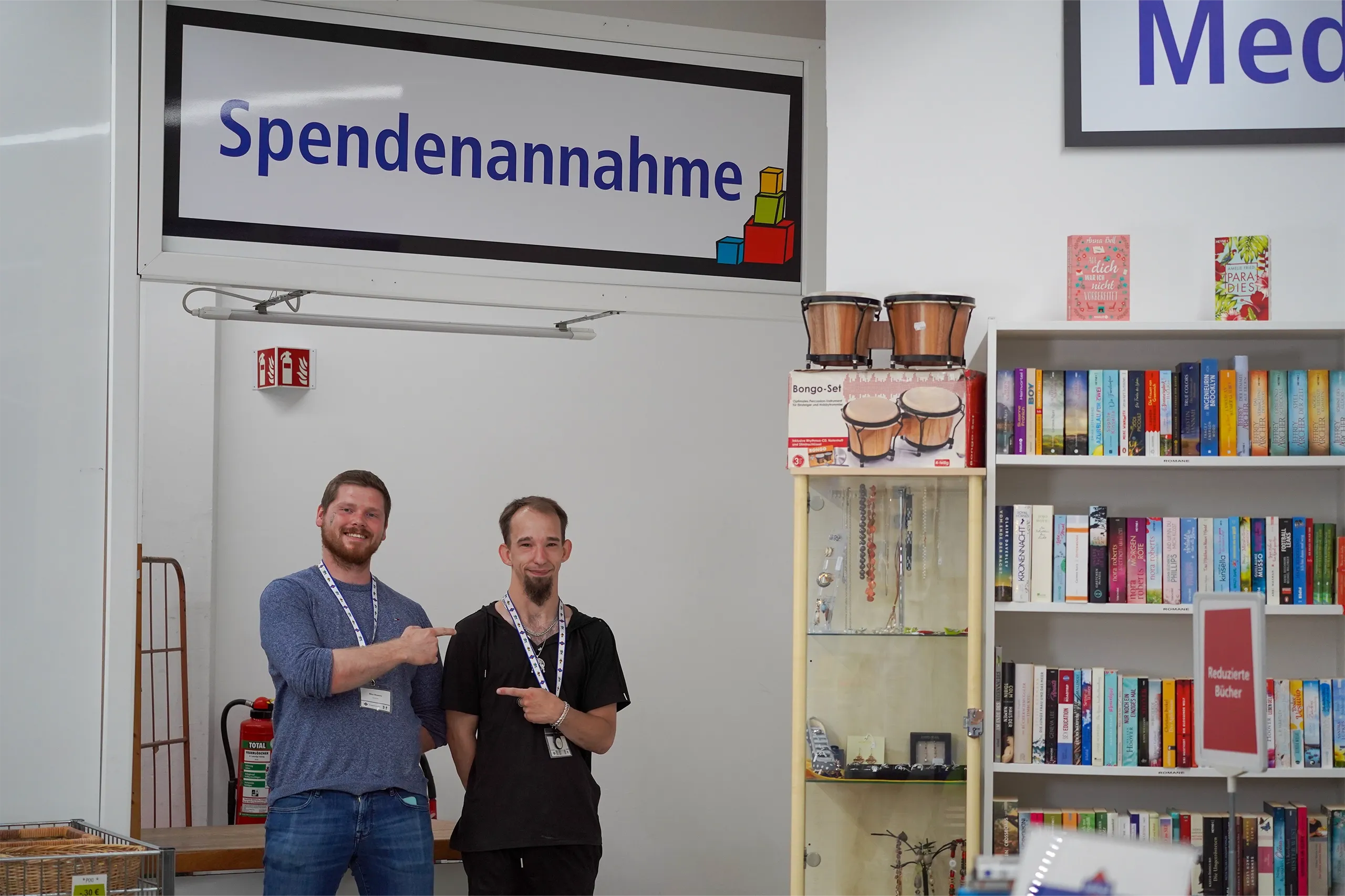 Zwei Personen stehen in einem Raum der Spendenannahme und zeigen aufeinander, während im Hintergrund Regale mit Büchern und Musikinstrumenten zu sehen sind.